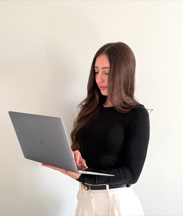 Eleni standing with a laptop against a warm off-white wall, working in natural light.